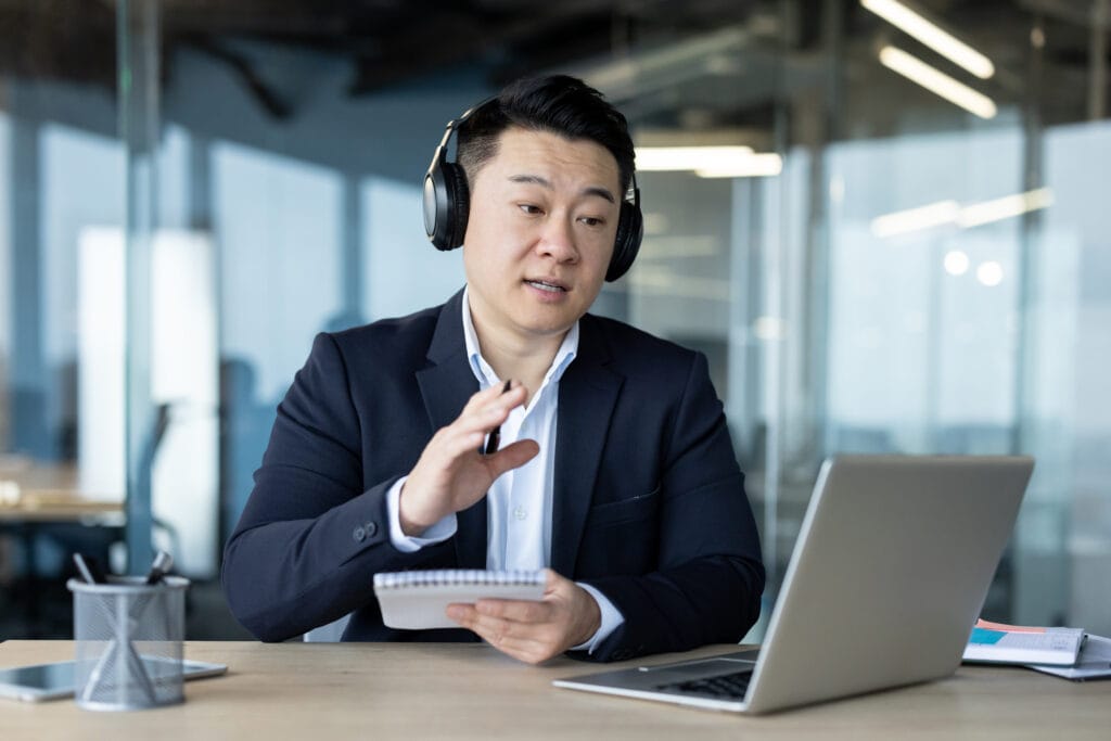 Asian young serious man in headphones and suit sitting in the office in front of a laptop, talking on a video call, taking notes